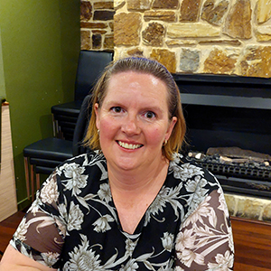 Jasmine is smiling. She is in front of a fireplace and wearing a blue floral patterned top