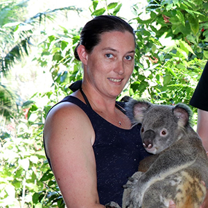Meegan is in a black singlet top, she is smiling at the camera and is holding a Koala. Behind her are green plants and trees.