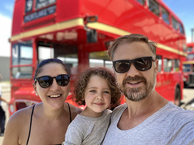 Angela is holding her son in her arms, and her husband is standing next to her. They are in front of a red London bus. he photo is taken after Angela has her stroke.