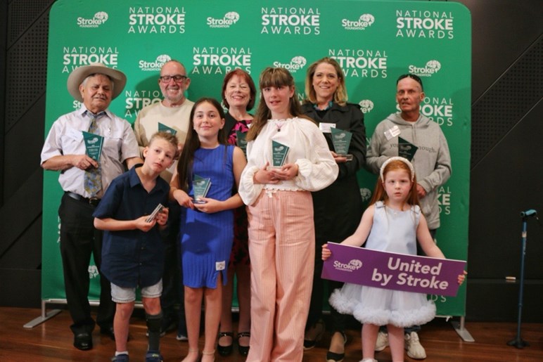 A group of award winners pose in front of a media wall with their awards